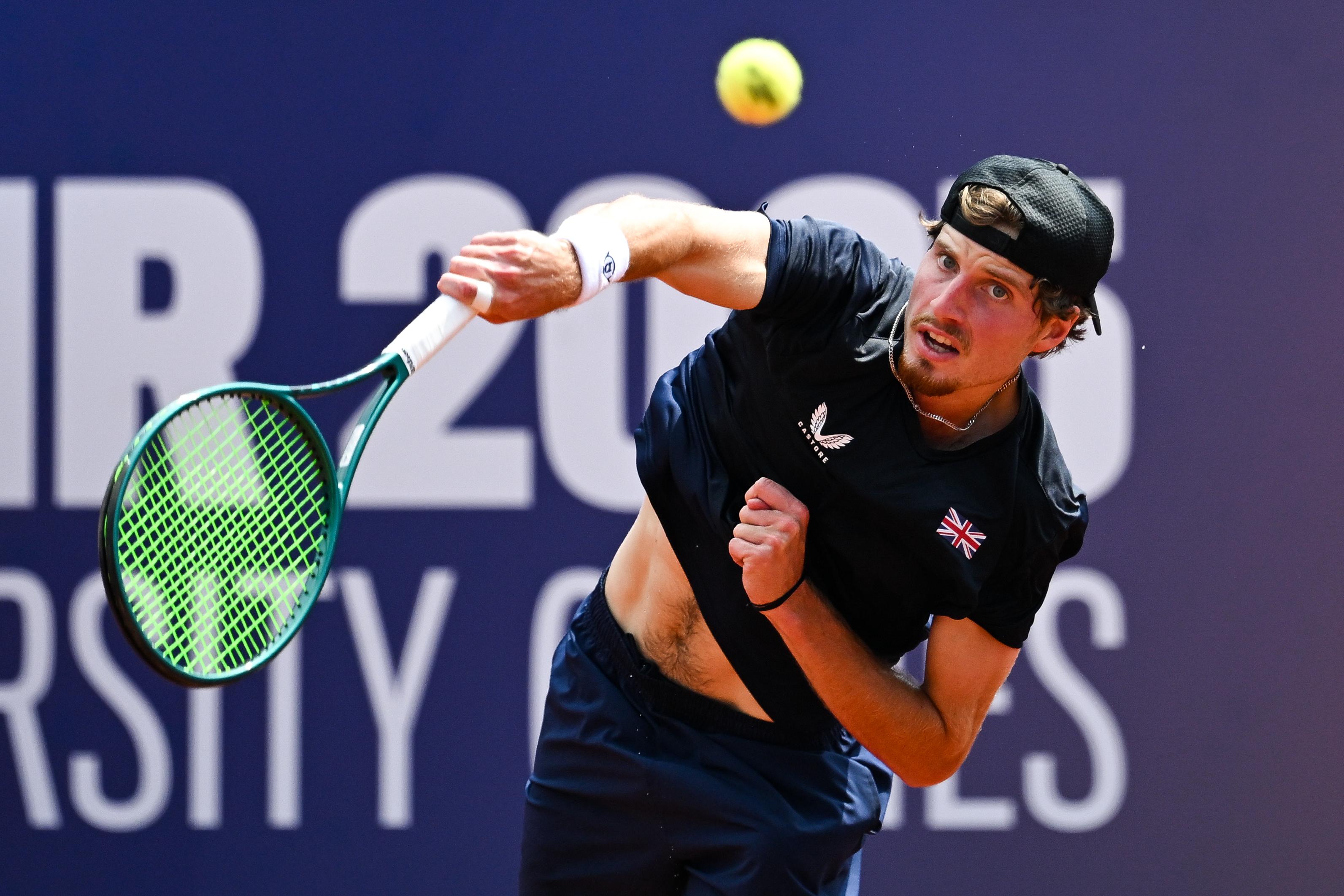 A man wearing a black cap and a dark blue shirt with the British flag on the left breast hits a tennis ball with a slightly strained expression on his face